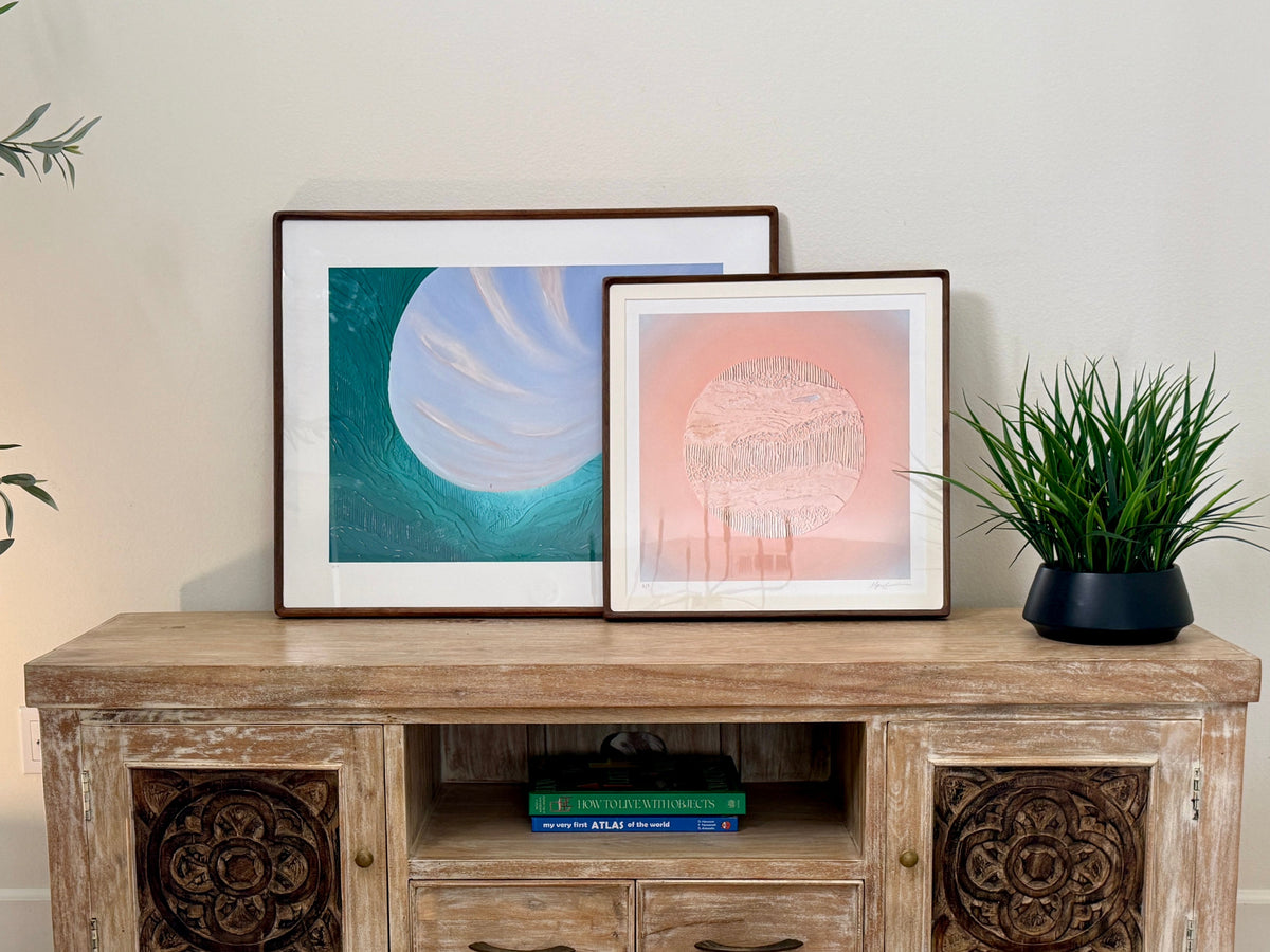A rustic light wood TV stand or console cabinet with carved doors and drawers, styled in a living room setting with two framed art prints, a potted plant, and books on top.